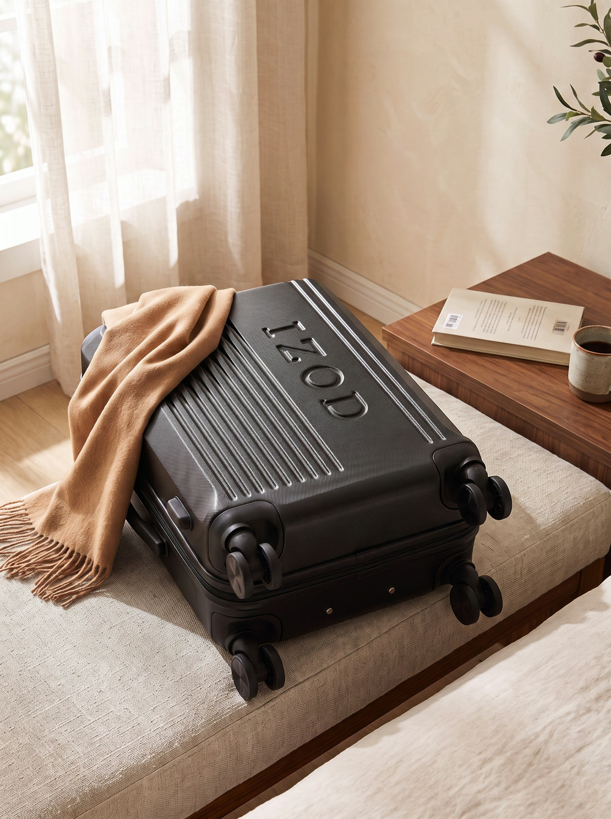 Black IZOD Zane luggage resting on a linen bench with a camel scarf and morning coffee