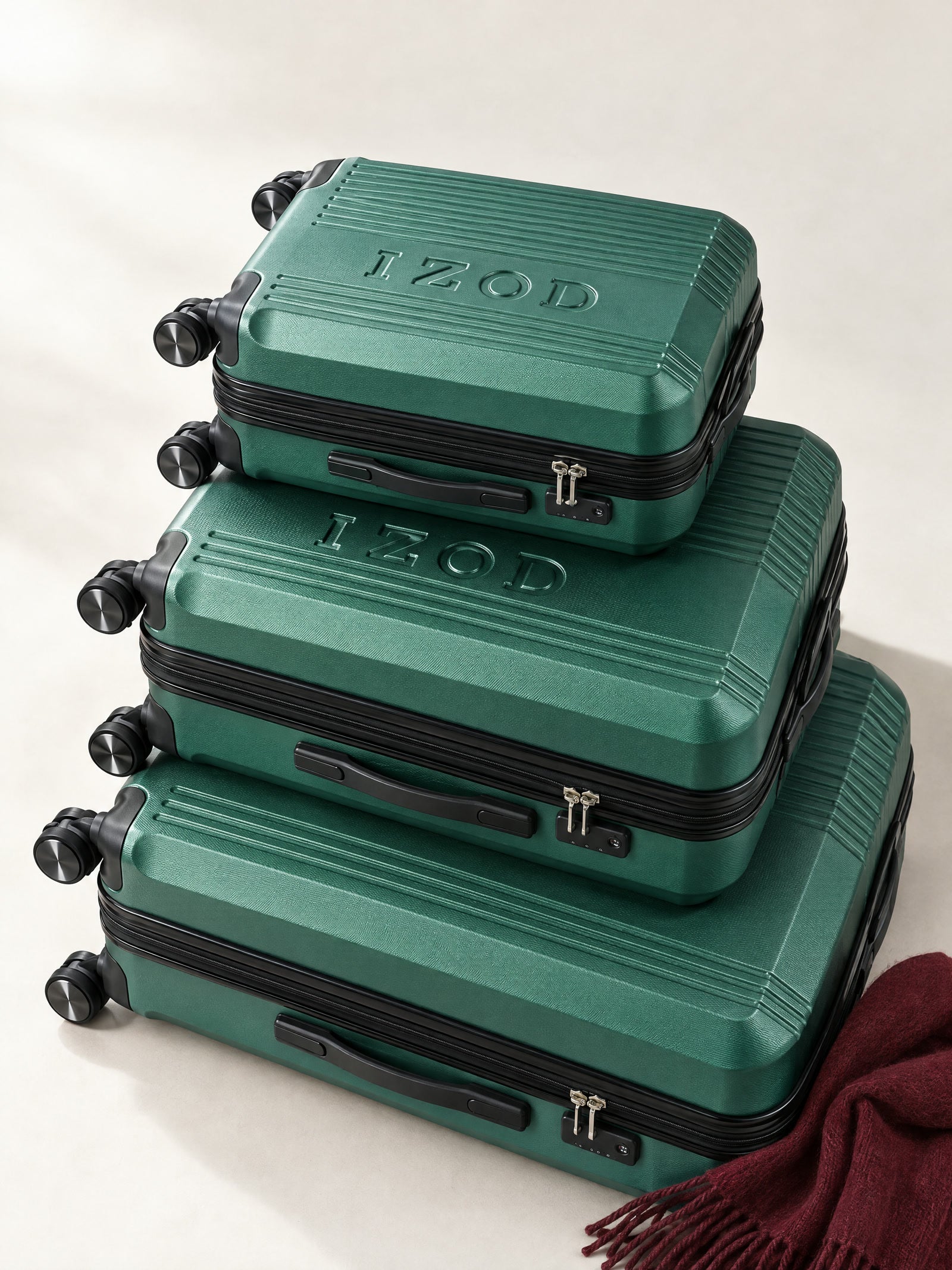 Three green IZOD Zane suitcases stacked on a warm studio floor with a burgundy scarf