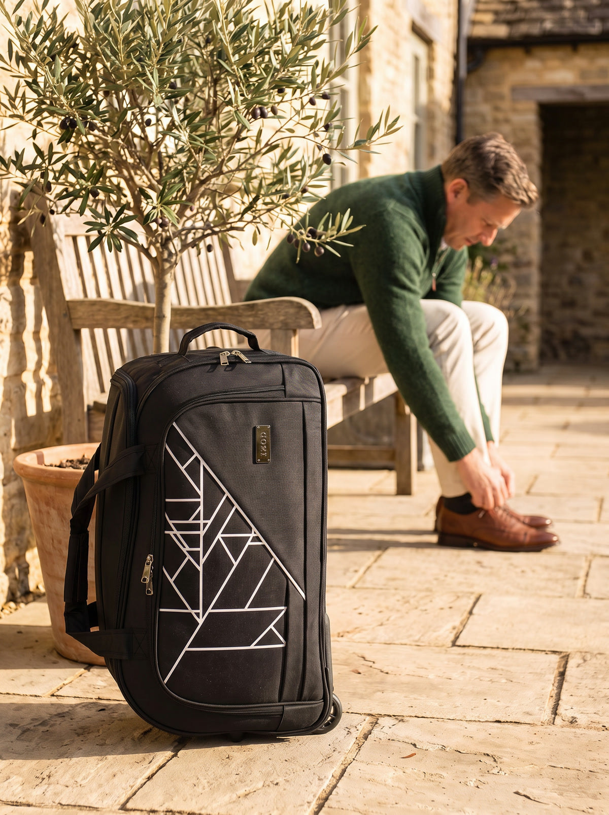 Black geometric-print IZOD rolling duffel beside a traveler getting ready on a stone terrace