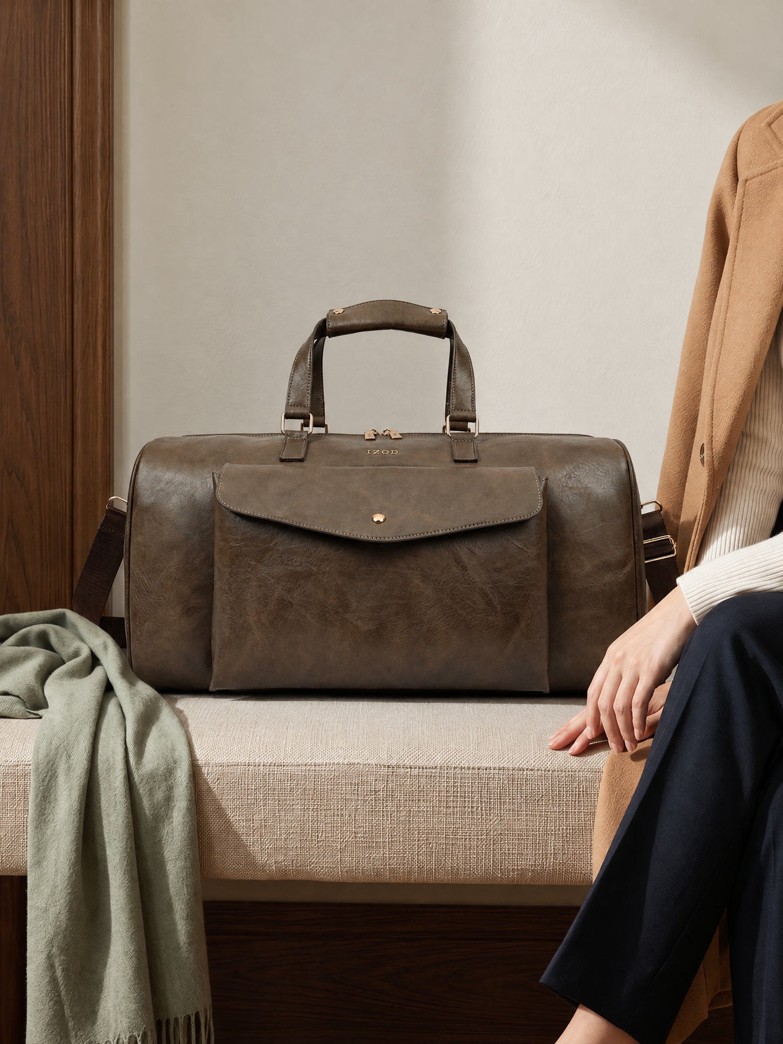The chocolate Atlas weekender beside a traveler on an upholstered bench, with a sage scarf