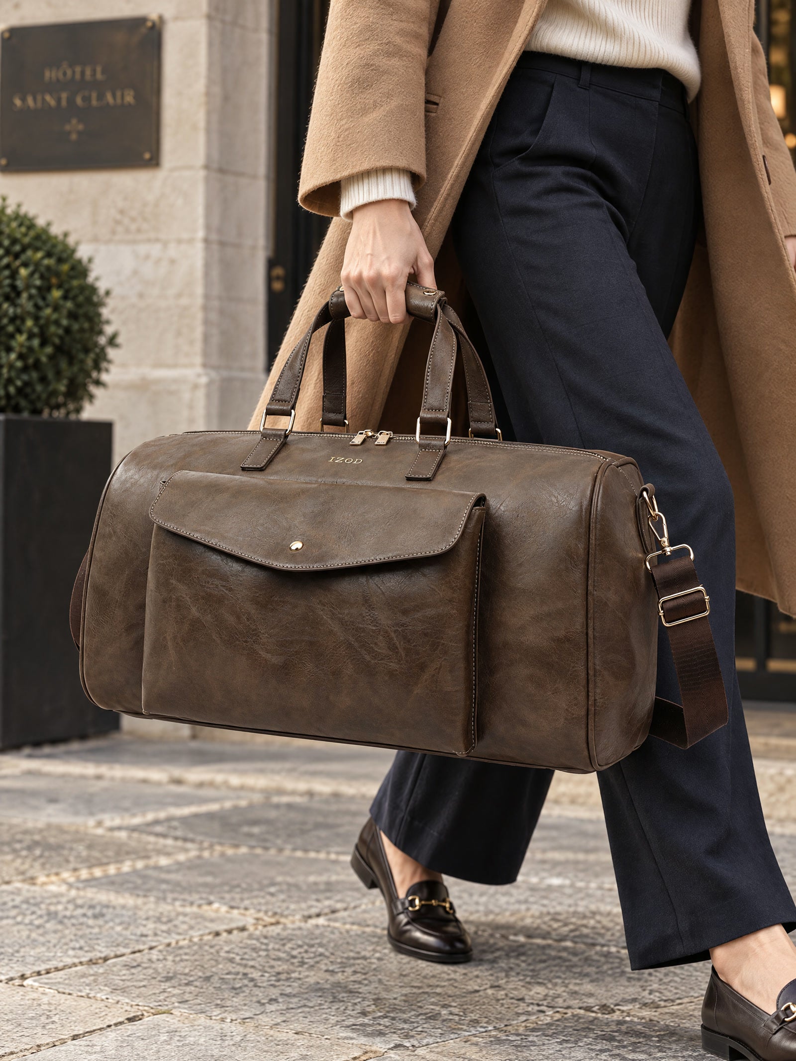 A traveler in a camel coat carrying the chocolate Atlas duffel outside a stone hotel